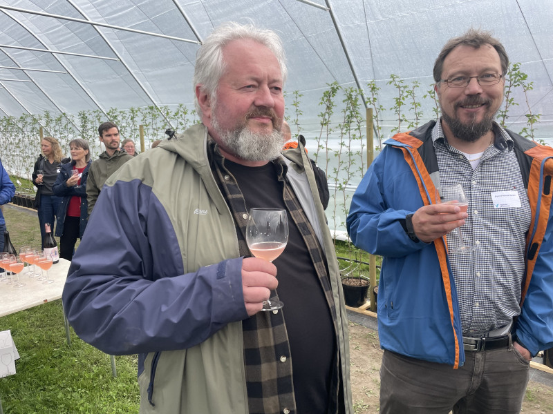 To middelaldrande menn med glas i handa står i plasttunnel og smiler.