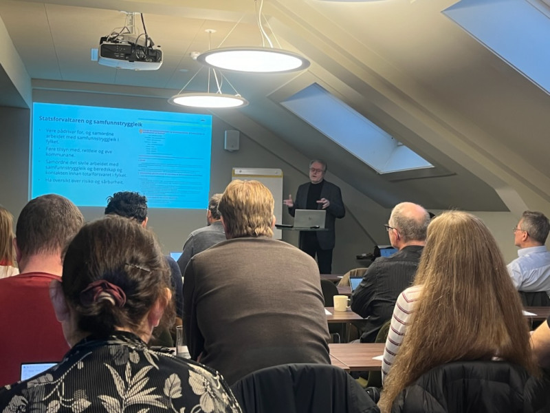 Møte i Bergen om KI-senter. Mange folk med ryggen til sit i eit møterom med skråtak. Ein person snakkar fremst i lokalet og viser ein presentasjon.