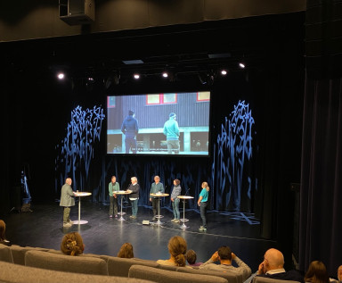 Scene med panel og utspørjar, lerret i bakgrunnen. Publikum i salen